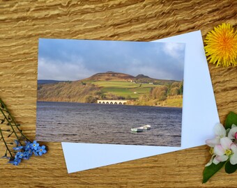 May include: A landscape photograph of a lake with a bridge and distant hills. Two small boats float on the water. A white card is angled in front of the photo, with a yellow flower and sprigs of blue and white flowers on a wooden surface.