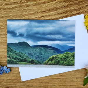 Op de afbeelding: Een kaart met een foto van een bergketen met een bewolkte lucht. De bergen zijn groen en de lucht is blauw met grijze wolken. De kaart ligt op een houten oppervlak met bloemen.