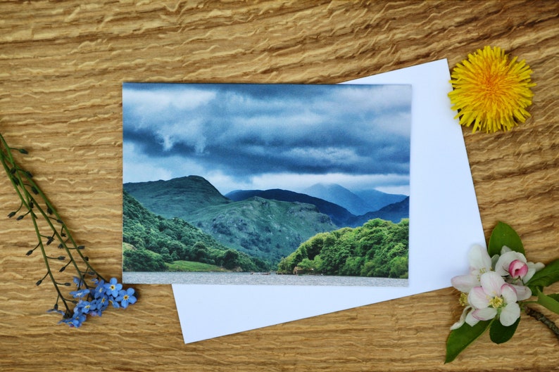 Op de afbeelding: Een kaart met een foto van een bergketen met een bewolkte lucht. De bergen zijn groen en de lucht is blauw met grijze wolken. De kaart ligt op een houten oppervlak met bloemen.