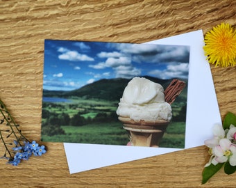 May include: A white ice cream cone with a flake topping, set against a scenic backdrop of a lake and mountains under a blue sky with white clouds.