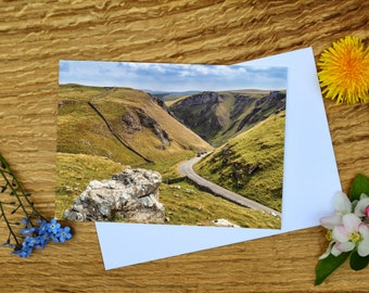 May include: A scenic photograph of a winding road through a green valley with a stone wall on the left side of the road. The road is surrounded by lush green hills and a blue sky with white clouds.