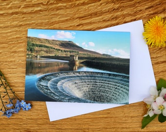 May include: A greeting card featuring a photograph of a dam with a large circular opening in the centre. The water is flowing through the opening, creating a swirling effect. The dam is located in a mountainous area with trees and a blue sky in the background.