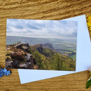Könnte beinhalten: Landschaftsfotografie eines Bergkamms mit grünen Bäumen und felsigen Klippen unter bewölktem Himmel. Eine weiße Karte im Vordergrund, mit einer gelben Blume und einem Zweig blauer Blumen und grünen Blättern.