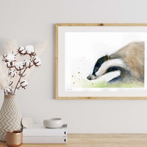 May include: Watercolor painting of a badger in a natural setting. The badger is brown and white with a black nose and is shown in profile. The background is a light green and white with splashes of gold paint.