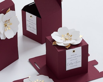 May include: Deep burgundy favor boxes featuring a white paper flower with gold accents on top. Each box includes a white rectangular tag with the text Thank you for being a part of our special day, Carolyn & Trance, and a date. Dimensions are 3.2x3.2x3.2 inches.