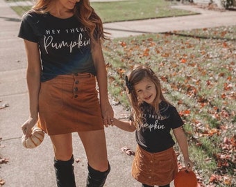 May include: A mother and daughter wearing matching black t-shirts with the text "Hey there Pumpkin" in white. They are both wearing brown corduroy skirts and holding pumpkins.