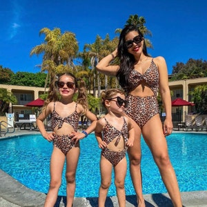Pu&ograve; includere: Tre persone con costumi da bagno leopardati abbinati e occhiali da sole posano vicino a una piscina. I costumi da bagno includono un costume intero e costumi da bagno a due pezzi. Palme e cielo blu sullo sfondo.