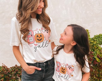 May include: Two people wearing white t-shirts with a pink and orange pumpkin, ghost, and bats graphic and the text "Boo yall". The people are wearing denim shorts and are looking at each other.