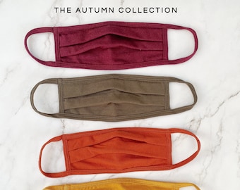 Könnte beinhalten: Fünf Stoffmasken in Herbstfarben: Burgunderrot, Olivgrün, Orange, Senfgelb und Braun. Die Masken sind gefaltet und haben elastische Ohrschlaufen. Der Text "THE AUTUMN COLLECTION" befindet sich oben im Bild.