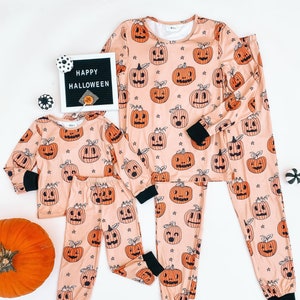 May include: A set of matching pajamas featuring a peach background with a repeating pattern of orange jack-o'-lantern faces. The pajamas come in two sizes, one for an adult and one for a child. The text "HAPPY HALLOWEEN" is visible on a black letter board in the background.