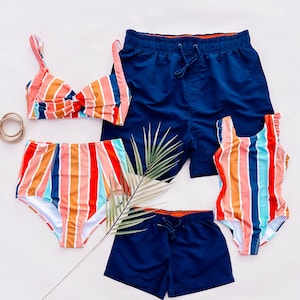 Può includere: Un set di quattro costumi da bagno a righe in uno schema di colori arcobaleno. Il set include un top bikini, uno slip bikini, un costume intero e pantaloncini da bagno. I costumi da bagno sono tutti blu navy con righe colorate.