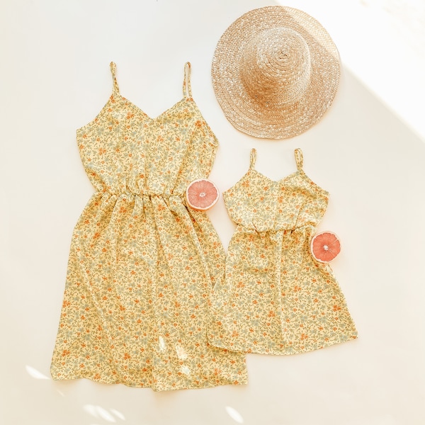 May include: Two yellow floral print sundresses with spaghetti straps. The larger dress has a cinched waist. A straw hat and two slices of grapefruit are also in the image.