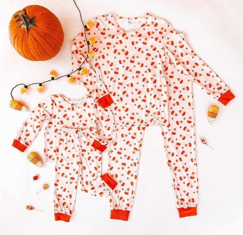 May include: A set of two matching pajamas with a candy corn print. The pajamas are white with orange and red candy corn designs. The adult pajamas have long sleeves and long pants. The child pajamas have long sleeves and long pants.