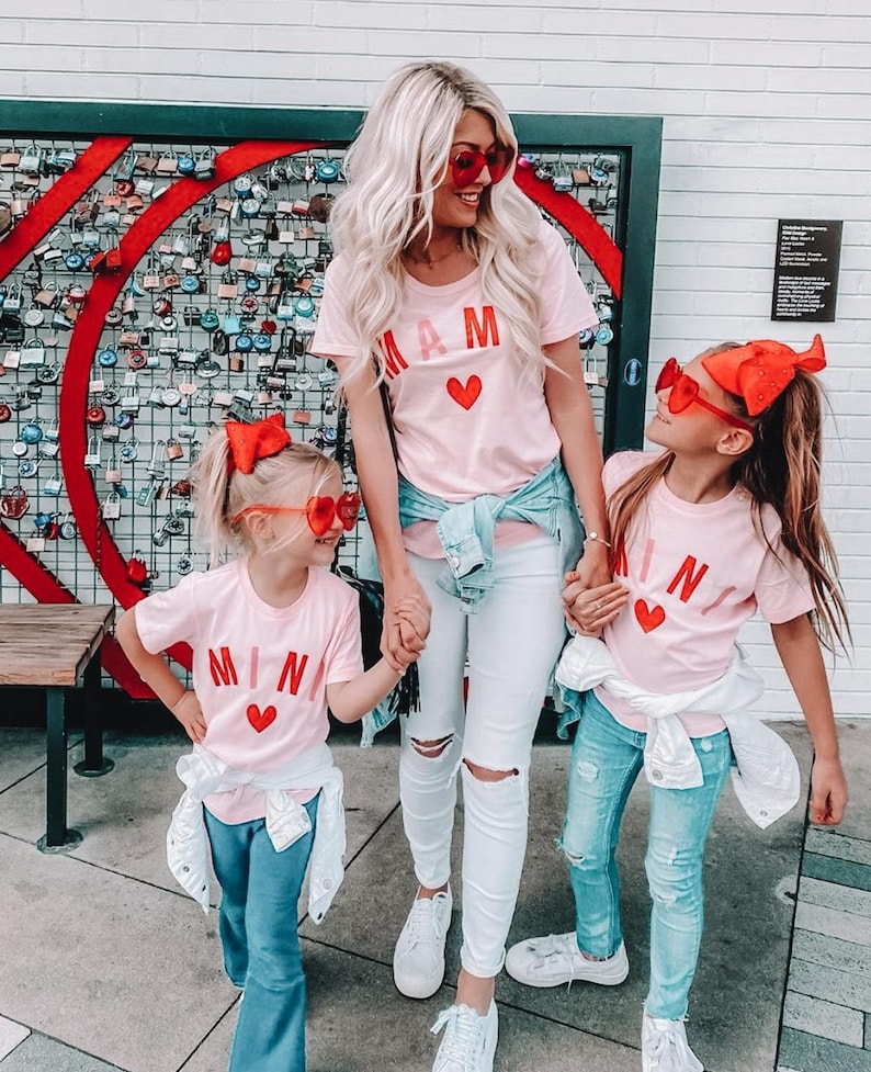 May include: A mother and her two daughters wearing pink t-shirts with the words "MAM" and "MINA" in red letters and a red heart. The mother is wearing white jeans and the daughters are wearing blue jeans. They are all wearing red heart-shaped sunglasses.