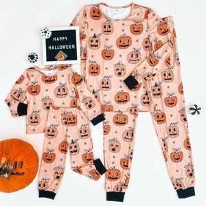 May include: A set of matching pajamas with a pumpkin print. The pajamas are peach colored with black outlines and black stars. The text "HAPPY HALLOWEEN" is on a black letter board in the background.