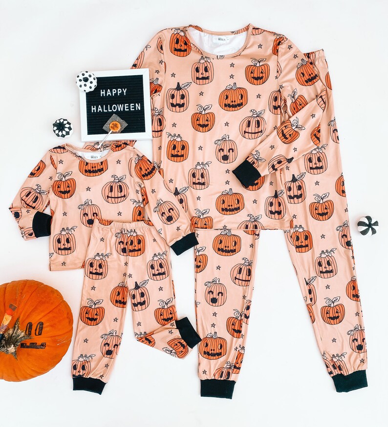 May include: A set of matching pajamas with a pumpkin print. The pajamas are peach colored with black outlines and black stars. The text "HAPPY HALLOWEEN" is on a black letter board in the background.