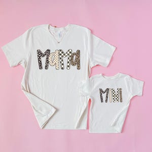 May include: Two off-white t-shirts with the words "Mama" and "Mini" printed on them. The word "Mama" is on the larger shirt and is designed with a mix of black and white polka dots, checkered, and leopard print patterns. The word "Mini" is on the smaller shirt.