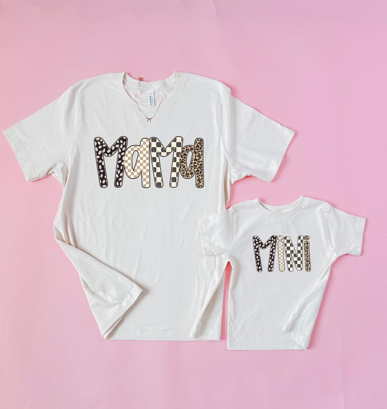 May include: Two off-white t-shirts with the words "Mama" and "Mini" printed on them. The word "Mama" is on the larger shirt and is designed with a mix of black and white polka dots, checkered, and leopard print patterns. The word "Mini" is on the smaller shirt.