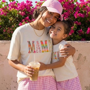 May include: Two people wearing matching cream-coloured t-shirts with the word "MAMA" in colourful letters. One person is wearing a pink baseball cap and holding a clear plastic cup with a straw. They are embracing in front of a pink flower bush.