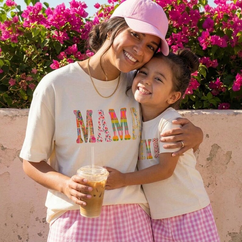 May include: Two people wearing matching cream-coloured t-shirts with the word "MAMA" in colourful letters. One person is wearing a pink baseball cap and holding a clear plastic cup with a straw. They are embracing in front of a pink flower bush.