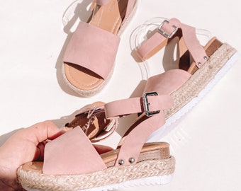 May include: A pair of pink platform sandals with a buckle closure and a jute sole. The sandals have a strap that goes around the ankle.