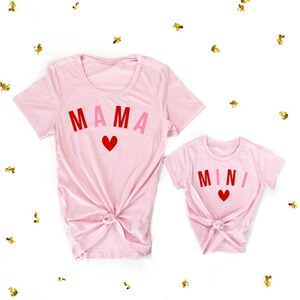 May include: Pink t-shirts with the words "MAMA" and "MINI" in red, pink, and white letters. A red heart is centered below the text on each shirt.