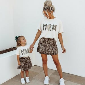 May include: A woman and a child wearing matching outfits. The woman wears a cream-colored t-shirt with the word "Mama" in a decorative font and a leopard print skirt. The child wears a similar outfit with the word "Mini".