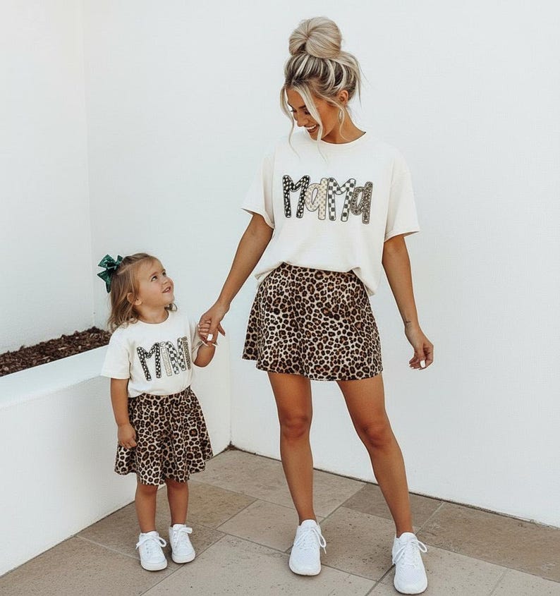 May include: A woman and a child wearing matching outfits. The woman wears a cream-colored t-shirt with the word "Mama" in a decorative font and a leopard print skirt. The child wears a similar outfit with the word "Mini".