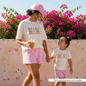 May include: Two people in matching outfits. One wears a cream t-shirt with the word "MAMA" in colourful letters, pink gingham shorts, and a pink cap. The other wears a cream t-shirt with the word "MINI" in colourful letters and pink gingham shorts.