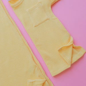 May include: A set of yellow waffle-knit clothing items, including a short-sleeved top with a pocket and a long, rectangular piece. The label "MIABELLA" is visible on the top. The items are displayed on a pink background.