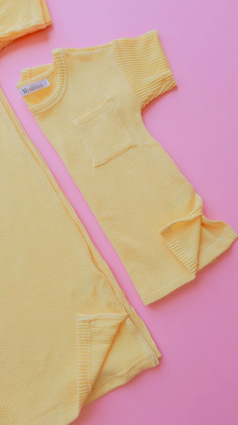 May include: A set of yellow waffle-knit clothing items, including a short-sleeved top with a pocket and a long, rectangular piece. The label "MIABELLA" is visible on the top. The items are displayed on a pink background.