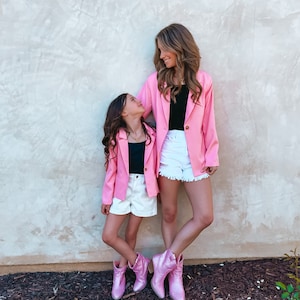 May include: Two people wearing pink blazers and white denim shorts stand in front of a textured wall. They are both wearing pink sparkly cowboy boots. The person at the back is smiling and looking at the person at the front. The person at the front is looking up at the person at the back.