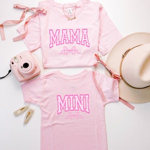 May include: Two pink t-shirts with the words "MAMA" and "MINI" in pink letters with a pink bow design. The shirts are on a white background.
