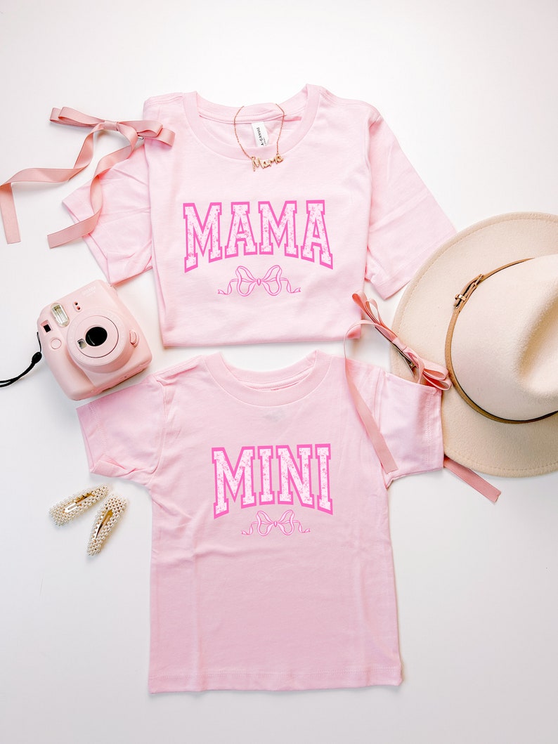 May include: Two pink t-shirts with the words "MAMA" and "MINI" in pink letters with a pink bow design. The shirts are on a white background.