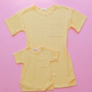 May include: Two yellow ribbed t-shirts, one adult and one child size, with short sleeves and a chest pocket. The shirts are on a pink background. The adult shirt has a longer length and side slits. The brand name is visible on the label.