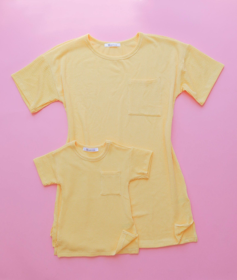 May include: Two yellow ribbed t-shirts, one adult and one child size, with short sleeves and a chest pocket. The shirts are on a pink background. The adult shirt has a longer length and side slits. The brand name is visible on the label.