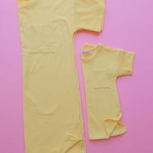 May include: Two yellow corduroy shirts, one adult-sized and one child-sized, are displayed on a pink background. Each shirt has short sleeves, a chest pocket, and a tag that reads "Mishella". The adult shirt is a long, tunic-style design.