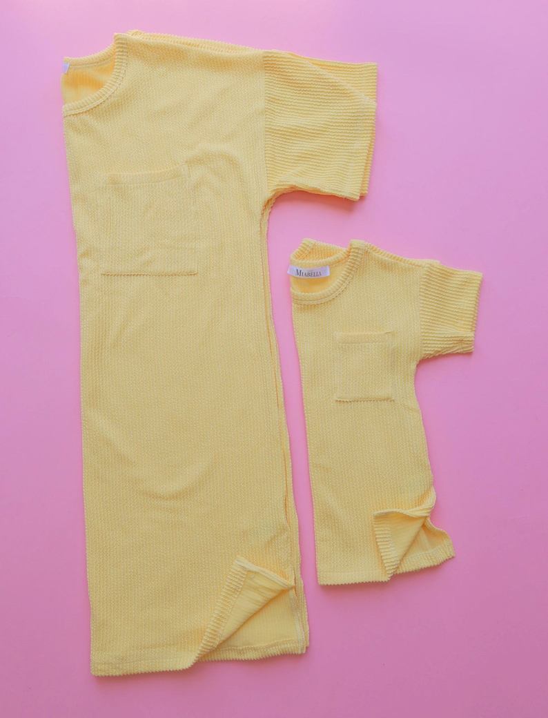 May include: Two yellow corduroy shirts, one adult-sized and one child-sized, are displayed on a pink background. Each shirt has short sleeves, a chest pocket, and a tag that reads "Mishella". The adult shirt is a long, tunic-style design.