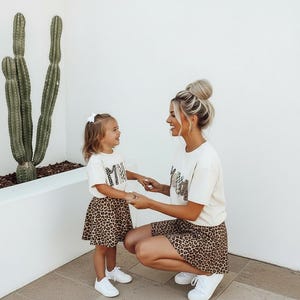 May include: Matching cream-colored t-shirts and leopard print skirts. The shirts have the word "MAMA" printed on them. The child is wearing a white bow and white sneakers. The adult is wearing white sneakers.