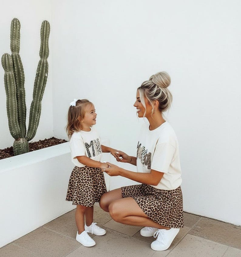 May include: Matching cream-colored t-shirts and leopard print skirts. The shirts have the word "MAMA" printed on them. The child is wearing a white bow and white sneakers. The adult is wearing white sneakers.