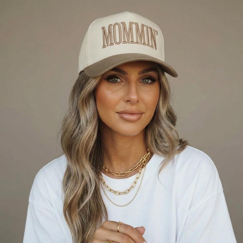 May include: A beige and brown baseball cap with the word "MOMMIN" embroidered on the front. The cap has a curved brim and a structured fit. The person in the image is wearing a white t-shirt and gold necklaces.