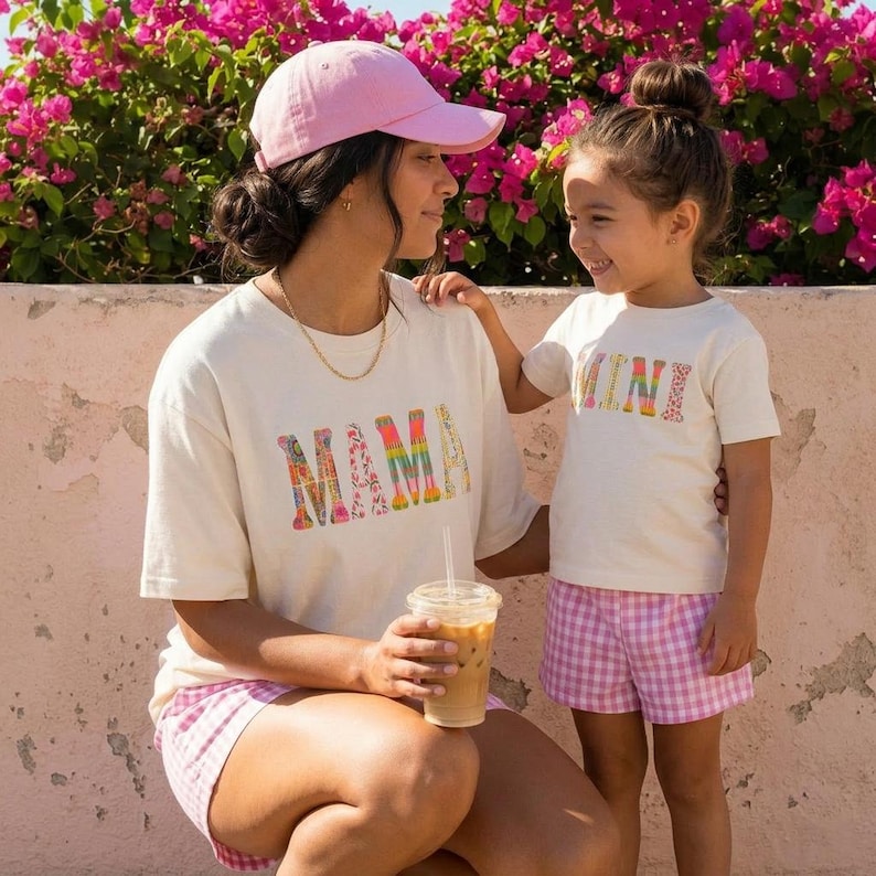 May include: Two cream-coloured t-shirts, one with "MAMA" and the other with "MINI" in patterned letters. The adult wears a pink cap and pink gingham shorts. The child wears pink gingham shorts. Pink flowers are in the background.