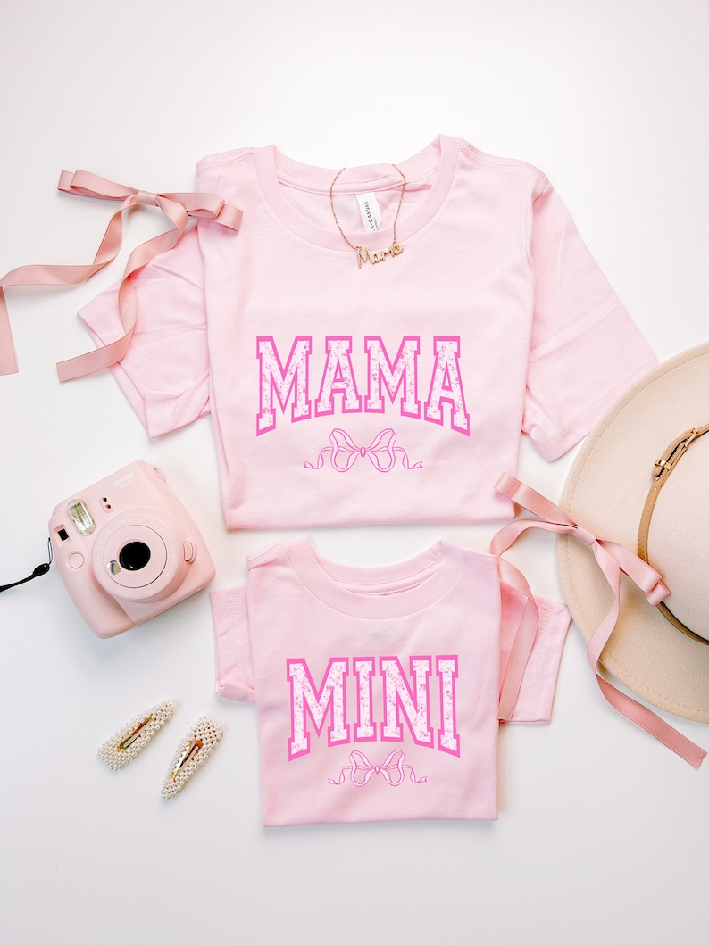 May include: Two pink t-shirts with the words "MAMA" and "MINI" in pink letters with white polka dots. The shirts are folded and laid out on a white surface with a pink ribbon, a pink camera, and a straw hat.