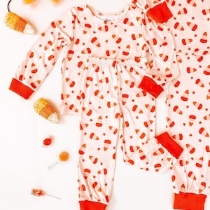 May include: A set of pajamas with a candy corn print. The pajamas are pink with orange and white candy corn designs and red trim.