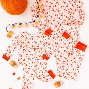 May include: A set of pink and orange pajamas with a candy corn print. The pajamas are on a white background with a pumpkin and string lights.