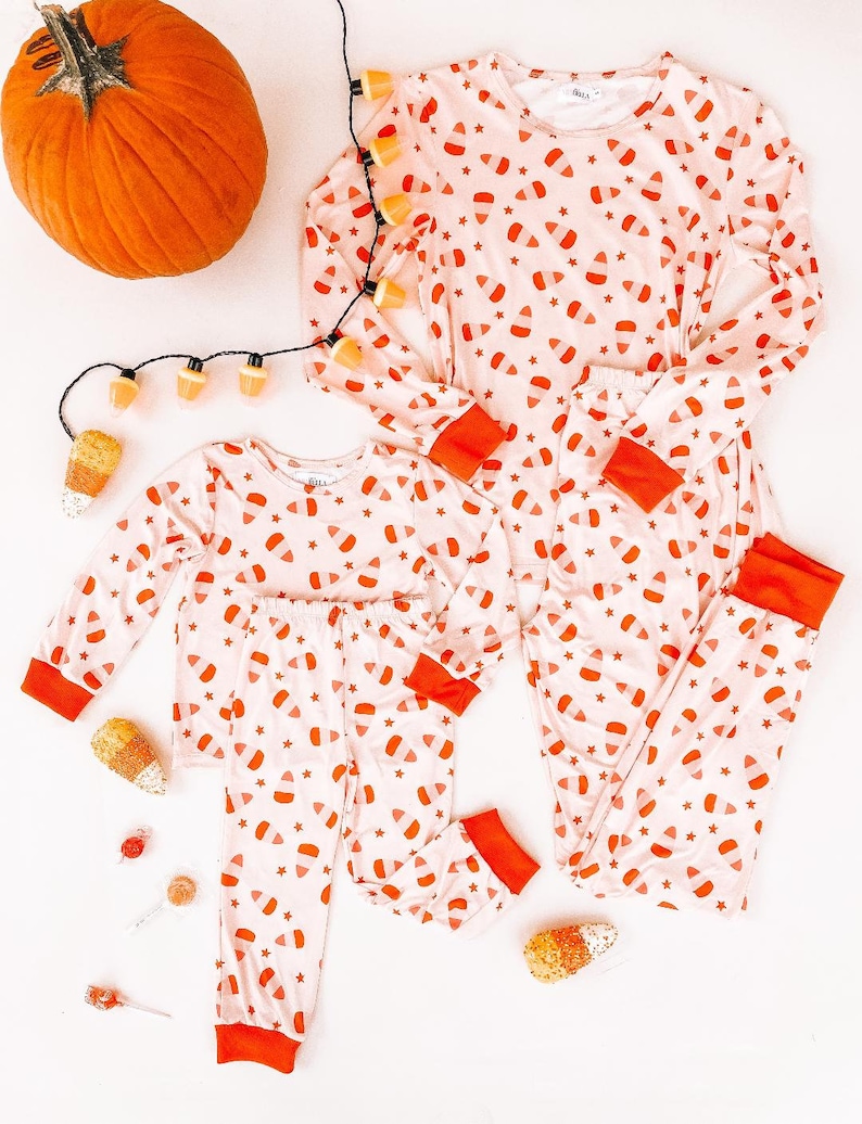 May include: A set of pink and orange pajamas with a candy corn print. The pajamas are on a white background with a pumpkin and string lights.