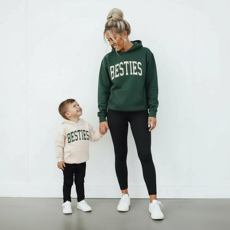 May include: A woman and a child wearing matching hoodies with the word "BESTIES" in white block letters. The woman wears a dark green hoodie, black leggings, and white sneakers. The child wears a beige hoodie, black pants, and white sneakers.