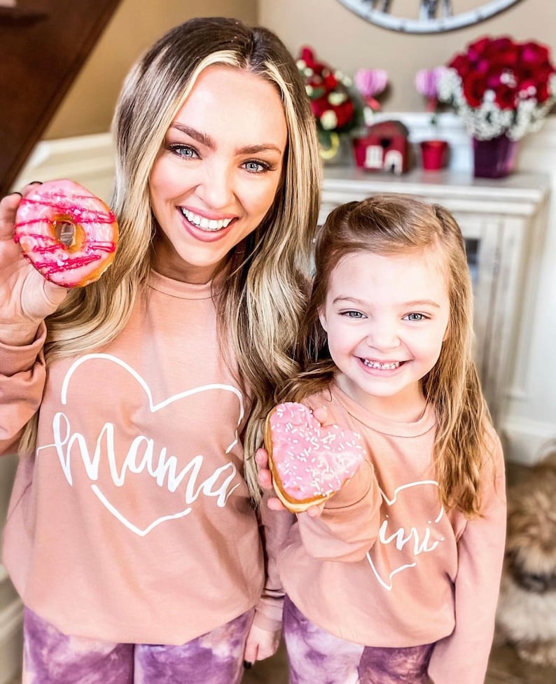 May include: A mother and daughter wearing matching pink sweatshirts with heart-shaped designs. The sweatshirts say "Mama" and "Mini" in white script. The mother is holding a pink frosted donut with sprinkles. The daughter is holding a pink heart-shaped donut with sprinkles.