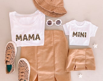 May include: A white t-shirt with the word "MAMA" printed in brown and black leopard print, a white t-shirt with the word "MINI" printed in brown and black leopard print, a brown skirt, a brown skirt, a pair of white sunglasses, a brown bucket hat, and a pair of brown and black leopard print sneakers.