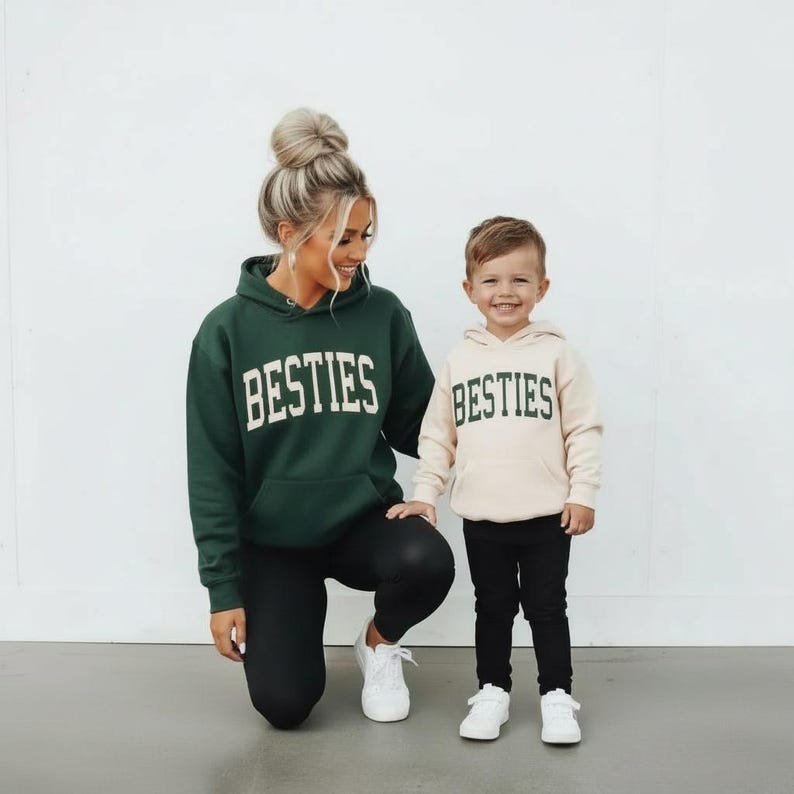 May include: A woman and a child wearing matching green and beige hoodies with the word "BESTIES" in white block letters. The woman is wearing black leggings and white sneakers. The child is wearing black pants and white sneakers.